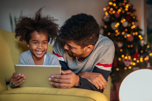 A parent is using a tablet to assist a child with an interactive educational app, both smiling and engaged in the activity. The setting is a well-lit and comfortable living room.