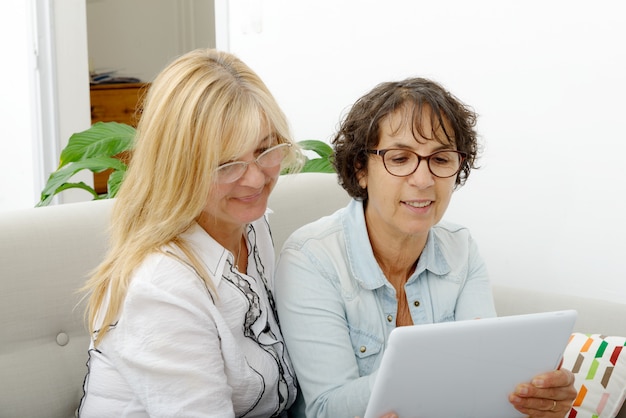 Two senior women smiling and looking at a tablet in a bright, modern living room. They are discussing finances, with charts and graphs visible on the screen.
