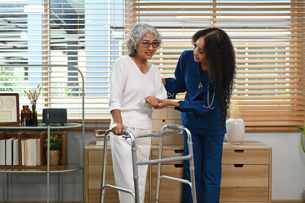 An elderly woman using a walker in her living room. Her daughter is assisting her and smiling. The environment is comfortable and supportive, indicating the availability of necessary care equipment.