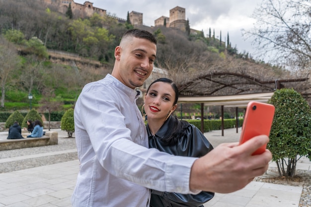 A group of Chinese tourists taking photos in front of the Alhambra in Granada, Spain. The tourists are smiling and interacting with the surroundings, showcasing their enjoyment of the experience. The Alhambra is prominently featured in the background, highlighting its historical and architectural significance.